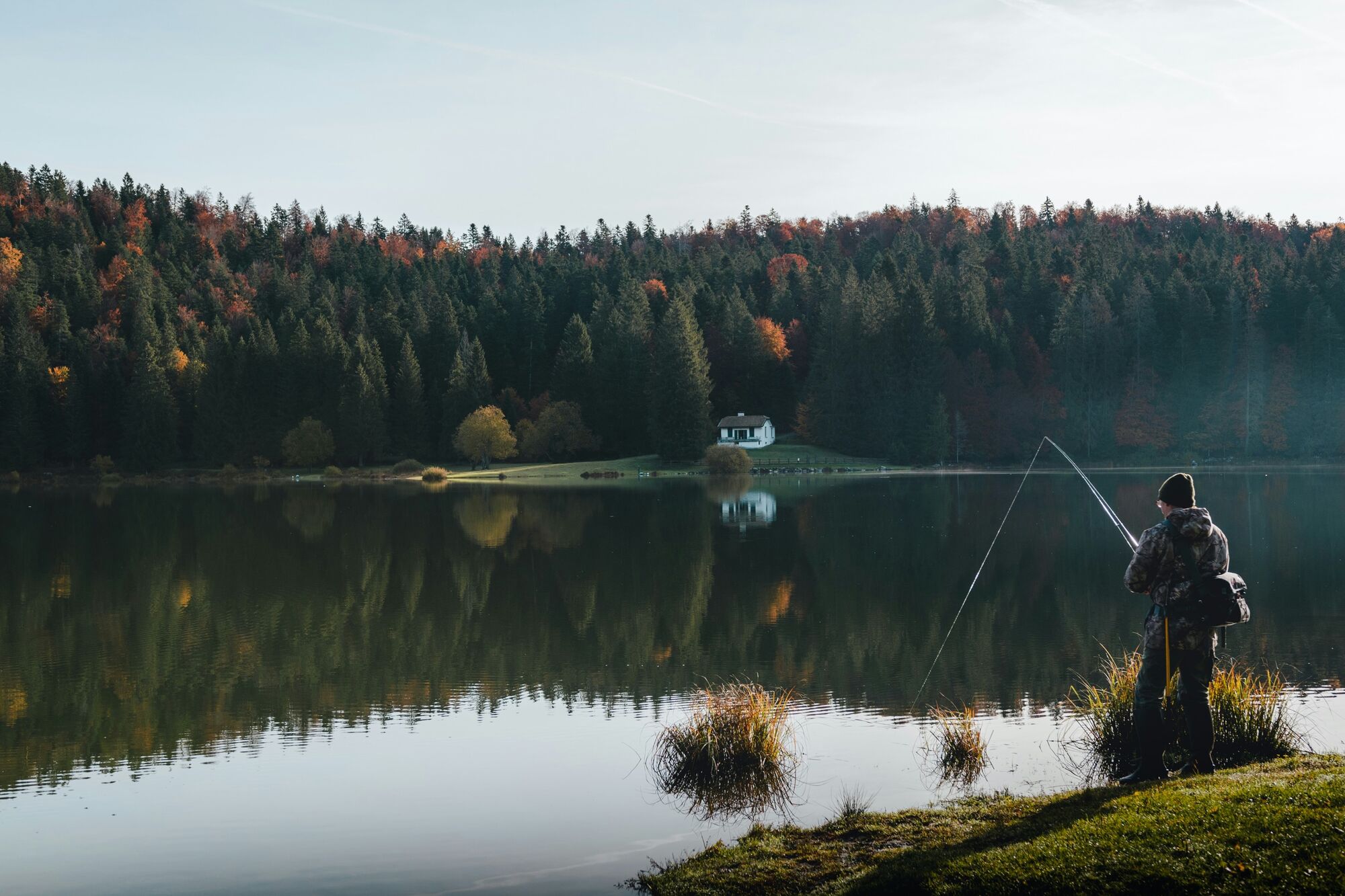 An angler fishing from the shore of a still lake ringed by autumn forest.